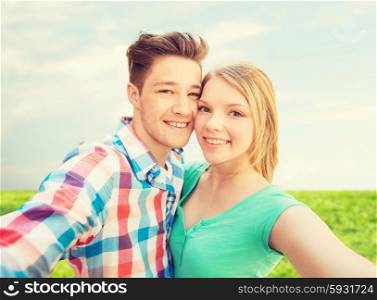 people, love, vacation, technology and summer concept - happy couple taking selfie with smartphone or camera over blue sky and grass background