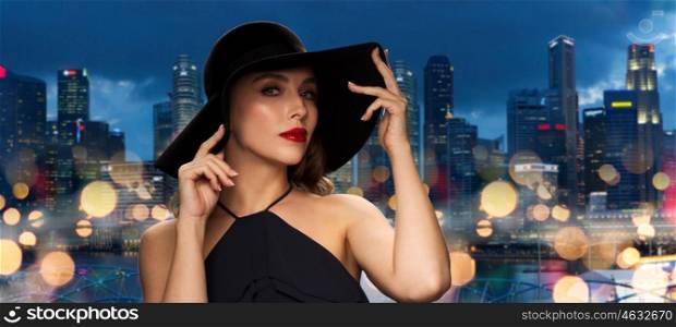 people, luxury and fashion concept - beautiful woman in black hat over nigh lights and singapore city skyscrapers background