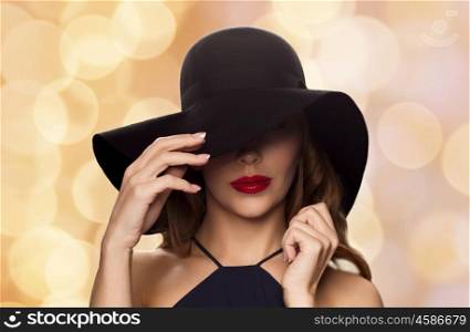 people, luxury and fashion concept - beautiful woman in black hat over holidays lights background