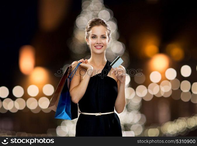 people, luxury and sale concept - beautiful woman with credit card and shopping bags over christmas tree lights background. woman with credit card and shopping bags