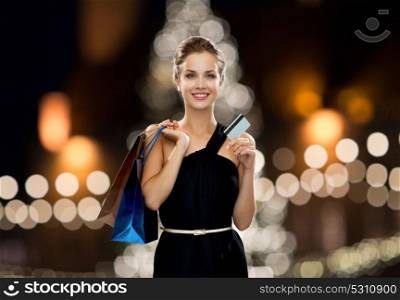 people, luxury and sale concept - beautiful woman with credit card and shopping bags over christmas tree lights background. woman with credit card and shopping bags