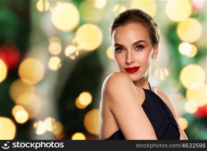 people, luxury, christmas, and fashion concept - beautiful woman in black with red lips over holidays lights background