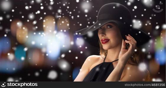 people, luxury, christmas, holidays and fashion concept - beautiful woman in black hat over blurred night lights background and snow