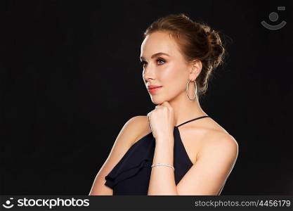 people, luxury, jewelry and fashion concept - beautiful woman in black wearing diamond earrings and bracelet over dark background