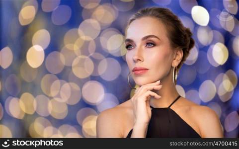 people, luxury, jewelry and fashion concept - beautiful woman in black wearing diamond earring and ring over blue holidays lights background
