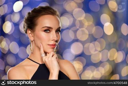 people, luxury, jewelry and fashion concept - beautiful woman in black wearing golden earrings and ring with diamonds over blue holidays lights background