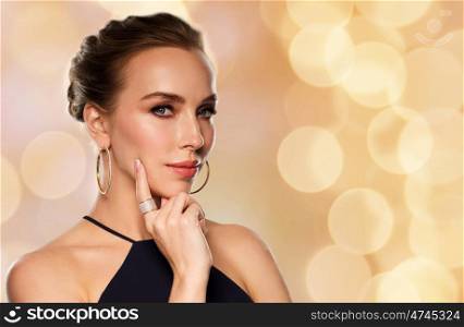 people, luxury, jewelry and fashion concept - beautiful woman in black wearing golden earrings and ring with diamonds over holidays lights background