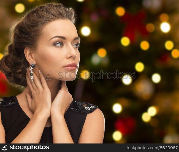 people, luxury, jewelry and fashion concept - beautiful woman in black wearing diamond earring over christmas tree lights background. beautiful woman with diamond jewelry on christmas