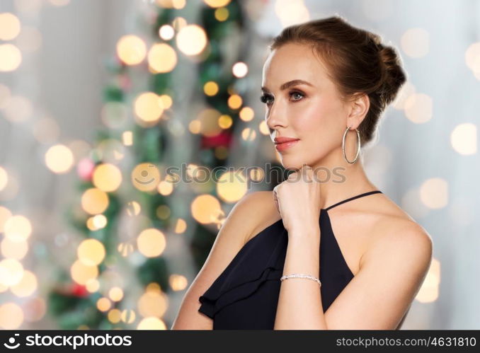 people, luxury, jewelry and holidays concept - beautiful woman in black wearing diamond earrings and bracelet over christmas tree lights background