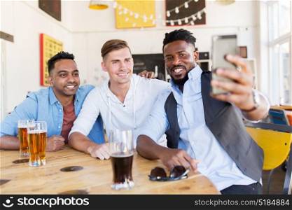 people, male friendship and technology concept - happy friends drinking beer and taking selfie by smartphone at bar or pub. friends taking selfie and drinking beer at bar