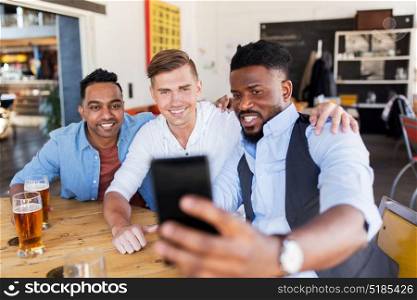 people, male friendship and technology concept - happy friends drinking beer and taking selfie by smartphone at bar or pub. friends taking selfie and drinking beer at bar