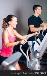People - man and woman - exercising in a gym on an elliptical trainer for more fitness