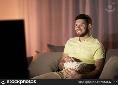 people, mass media, television and entertainment concept - happy smiling young man watching tv and eating popcorn at night at home. happy man with popcorn watching tv at night