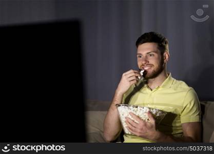 people, mass media, television and entertainment concept - happy smiling young man watching tv and eating popcorn at night at home. happy man with popcorn watching tv at night