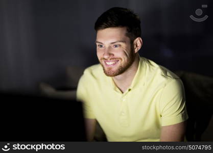 people, mass media, television and entertainment concept - happy smiling young man watching tv at night at home. happy smiling young man watching tv at night