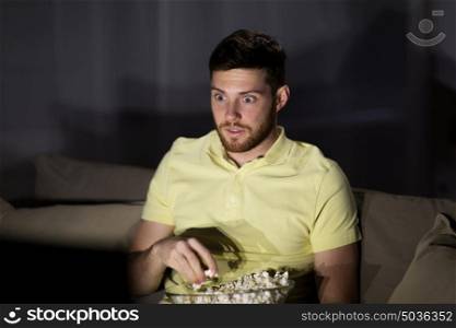 people, mass media, television and entertainment concept - scared young man watching tv and eating popcorn at night at home. man watching tv and eating popcorn at night
