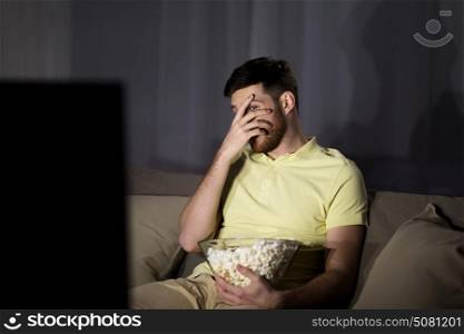 people, mass media, television and entertainment concept - scared young man watching tv and eating popcorn at night at home. scared man watching tv and eating popcorn at night