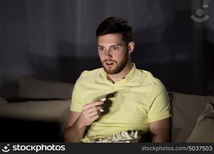 people, mass media, television and entertainment concept - young man watching tv and eating popcorn at night at home. man watching tv and eating popcorn at night