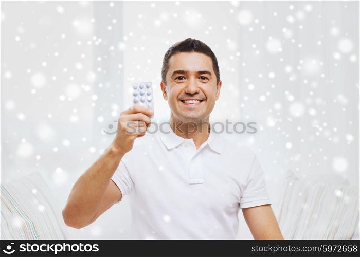 people, medication, medicine and health care concept - happy man showing pack of pills at home over snow effect