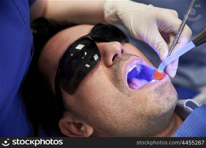 people, medicine, stomatology and health care concept - close up of dentist hand with dental curing light and mirror treating male patient teeth at dental clinic office