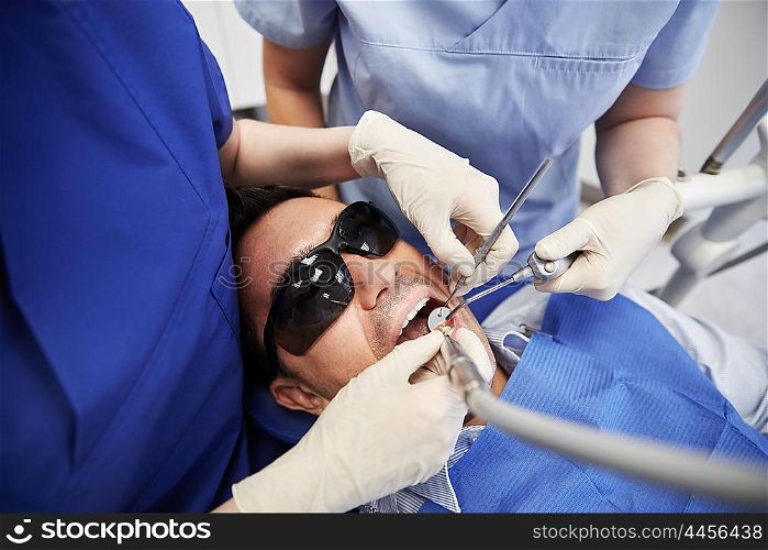 people, medicine, stomatology and health care concept - close up of dentist and assistant hands with dental mirror, drill and air water gun spray treating male patient teeth at dental clinic