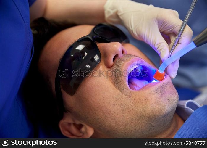 people, medicine, stomatology and health care concept - close up of dentist hand with dental curing light and mirror treating male patient teeth at dental clinic office