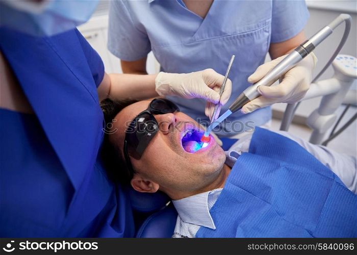 people, medicine, stomatology and health care concept - female dentist and assistant with dental curing light and mirror treating male patient teeth at dental clinic office