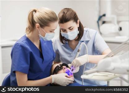 people, medicine, stomatology and health care concept - female dentists with dental curing light and mirror treating patient girl teeth at dental clinic office