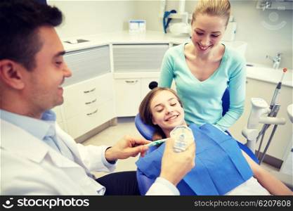 people, medicine, stomatology and health care concept - happy male dentist with toothbrush and jaw layout showing how to brush teeth to patient girl and her mother at dental clinic office