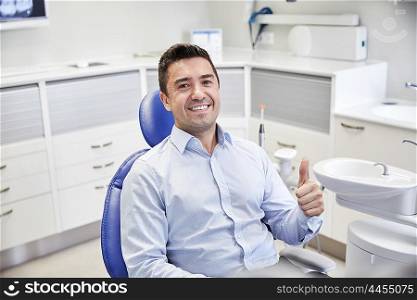 people, medicine, stomatology and health care concept - happy male patient sitting on dental chair and showing thumbs up at clinic office