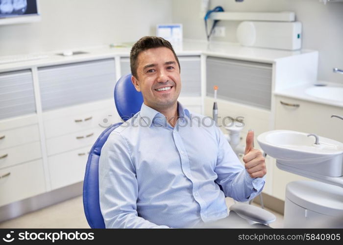 people, medicine, stomatology and health care concept - happy male patient sitting on dental chair and showing thumbs up at clinic office