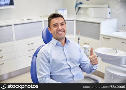 people, medicine, stomatology and health care concept - happy male patient sitting on dental chair and showing thumbs up at clinic office