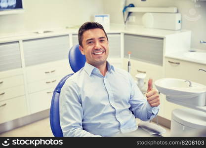 people, medicine, stomatology and health care concept - happy male patient sitting on dental chair and showing thumbs up at clinic office