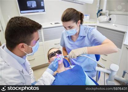 people, medicine, stomatology and health care concept - male dentist and assistant with dental curing light and mirror treating female patient teeth at dental clinic office