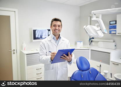 people, medicine, stomatology and healthcare concept - happy middle aged male dentist in white coat writing to clipboard at dental clinic office