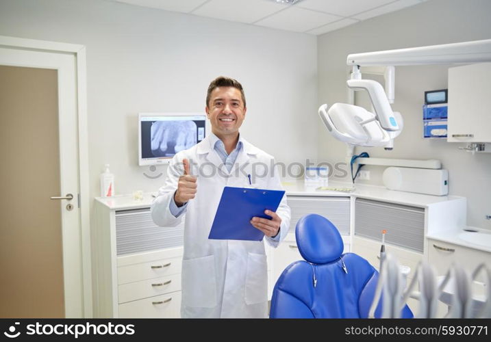 people, medicine, stomatology and healthcare concept - happy middle aged male dentist in white coat with clipboard showing thumbs up at dental clinic office