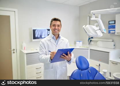 people, medicine, stomatology and healthcare concept - happy middle aged male dentist in white coat writing to clipboard at dental clinic office