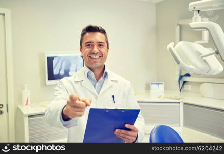 people, medicine, stomatology and healthcare concept - happy middle aged male dentist in white coat at with clipboard pointing finger to you at dental clinic office