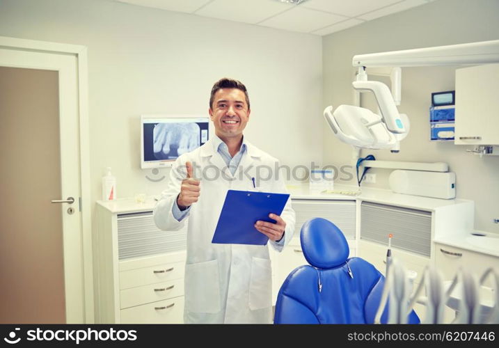 people, medicine, stomatology and healthcare concept - happy middle aged male dentist in white coat with clipboard showing thumbs up at dental clinic office