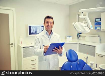 people, medicine, stomatology and healthcare concept - happy middle aged male dentist in white coat writing to clipboard at dental clinic office
