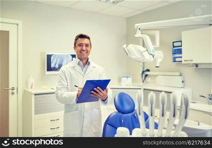 people, medicine, stomatology and healthcare concept - happy middle aged male dentist in white coat writing to clipboard at dental clinic office