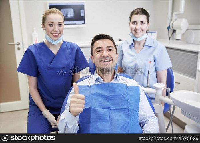 people, medicine, stomatology, gesture and health care concept - happy female dentist with assistant and man patient showing thumbs up at dental clinic office