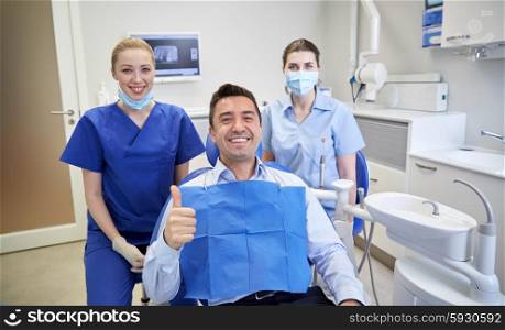people, medicine, stomatology, gesture and health care concept - happy female dentist with assistant and man patient showing thumbs up at dental clinic office