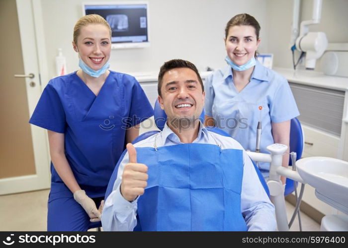 people, medicine, stomatology, gesture and health care concept - happy female dentist with assistant and man patient showing thumbs up at dental clinic office