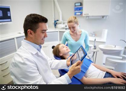 people, medicine, stomatology, technology and health care concept - happy male dentist looking at teeth x-ray on tablet pc computer and patient girl with her mother at dental clinic office