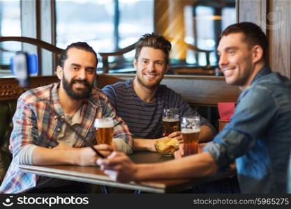 people, men, leisure, friendship and technology concept - happy male friends drinking beer and taking picture with smartphone selfie stick at bar or pub