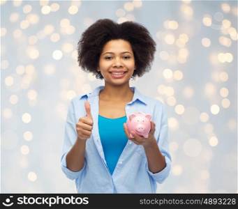 people, money saving and finances concept - happy african american young woman with piggy bank showing thumbs up over holidays lights background