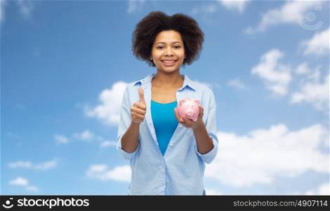 people, money saving and finances concept - happy african american young woman with piggy bank showing thumbs up over blue sky and clouds background. happy woman with piggy bank showing thumbs up