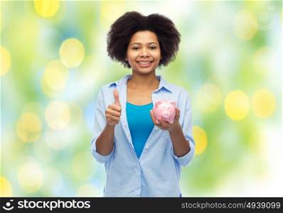people, money saving and finances concept - happy african american young woman with piggy bank showing thumbs up over summer green lights background. happy woman with piggy bank showing thumbs up