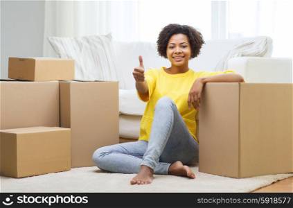 people, moving new place, gesture and repair concept - happy african american young woman with many cardboard boxes sitting on floor and showing thumbs up at home